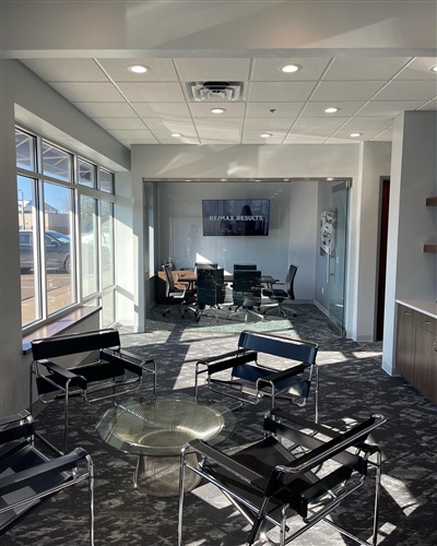 Modern office space with large windows, a glass table with black chairs in the foreground, and a conference room with a TV in the background.