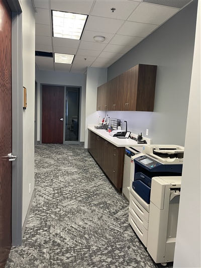 Office hallway with patterned carpet, wooden cabinets along one side, a copier machine, and a door leading to another room at the end.