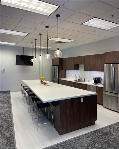 Modern office kitchen with a long island, barstools, pendant lights, stainless steel appliances, and wooden cabinets.