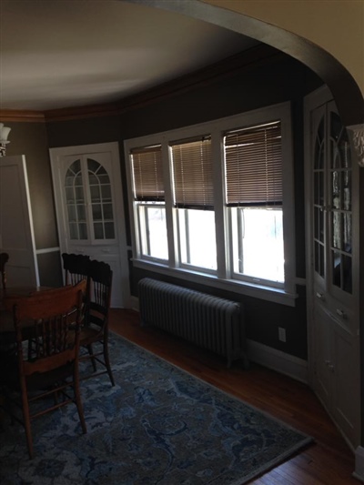 A dining room with dark walls, a wooden dining table and chairs, a large window with blinds, a blue rug, and an arched doorway.