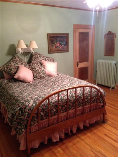 A bedroom with light green walls, a wooden floor, a bed with a floral bedspread and pink pillows, a wooden door, and a wall-mounted light fixture.