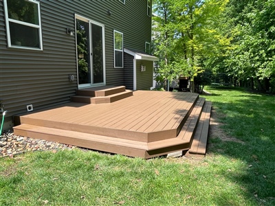 A backyard wooden deck attached to a house, featuring steps and surrounded by a well-maintained lawn with trees in the background.