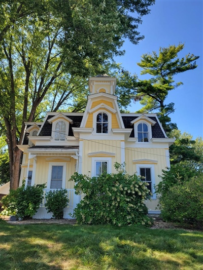 A charming two-story yellow house with white trim, multiple dormer windows, and a tall central tower, surrounded by lush greenery and trees.