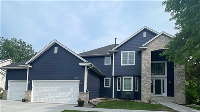 A modern two-story house with a dark blue exterior, white trim, and stone accents, featuring a large three-car garage and a well-maintained driveway.