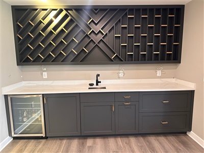 A modern kitchen countertop with dark cabinets, a built-in mini fridge, a sink, and a decorative geometric wine rack above.