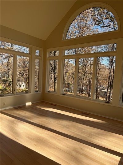 A sunlit room with large arched and rectangular windows overlooking a wooded area, featuring light wood floors.