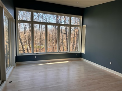 A room with large windows offering a view of a wooded area, featuring dark blue walls and light wood floors.