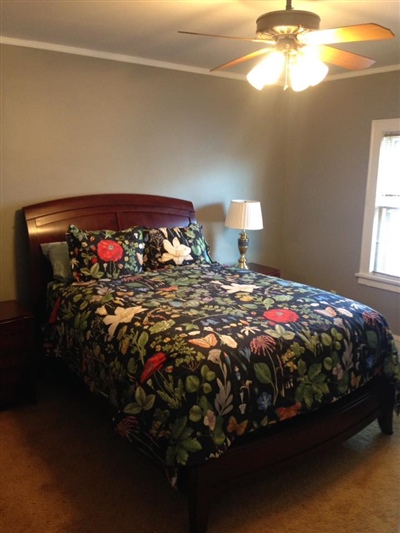 A bedroom with gray walls, a bed with a colorful floral bedspread, a ceiling fan, a nightstand with a lamp, and a window letting in natural light.