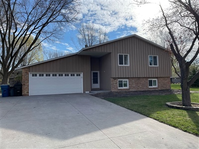 A two-story house with a brown exterior, white garage door, and a well-maintained lawn, surrounded by trees on a sunny day.