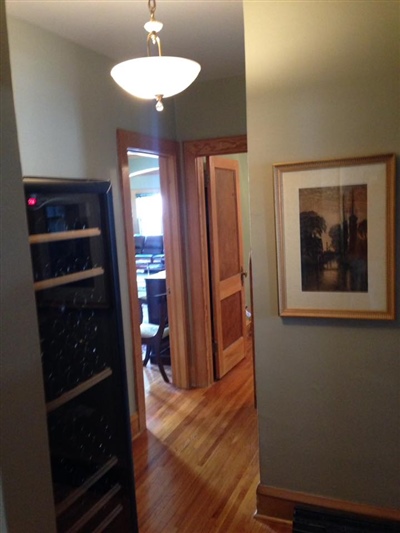 A hallway with light green walls, a framed picture, a wine rack, wooden doors, and a hanging light fixture.