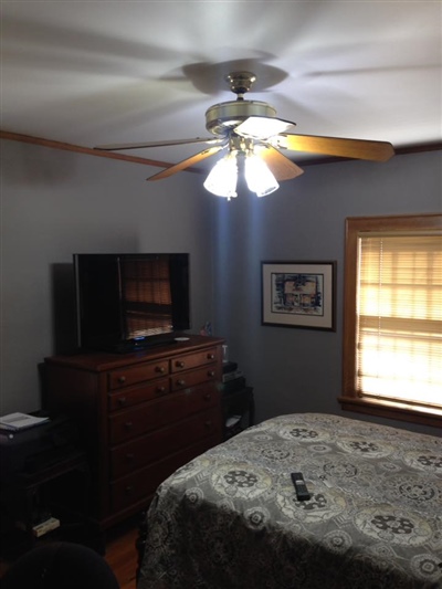 A bedroom with gray walls, a ceiling fan with lights, a bed with a patterned bedspread, a wooden dresser with a TV on top, and a window with blinds.