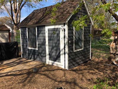 A small gray shed with white trim, surrounded by trees and located in a backyard.