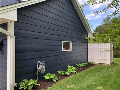 A side view of a house with dark blue siding, a small window, and a white fence, with neatly planted greenery along the foundation.