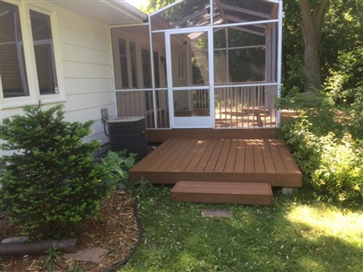 A backyard with a small wooden deck leading to a screened-in porch attached to a white house, surrounded by greenery.