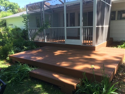 A backyard with a wooden deck and steps leading to a screened-in porch attached to a white house, surrounded by plants and greenery.