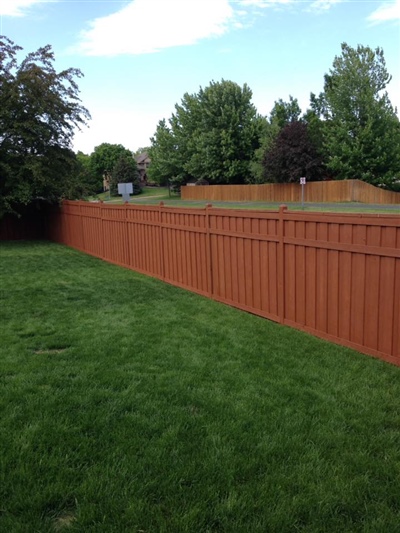 A backyard with a green lawn, bordered by a tall red wooden fence, and trees in the background.