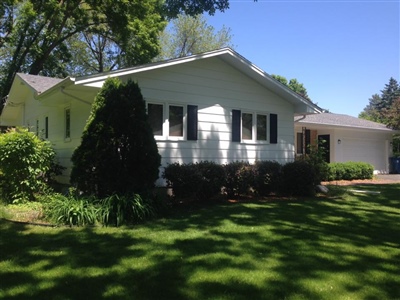 A white single-story house with a well-maintained lawn, shrubs, and trees, set in a sunny yard.