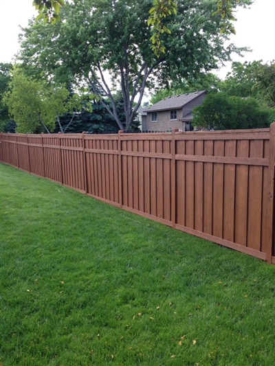 A backyard with a lush green lawn, bordered by a tall red wooden fence, with trees and a house visible in the background.