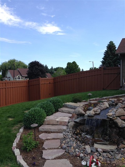 A backyard with a stone path leading to a water feature, surrounded by shrubs and greenery, and bordered by a red wooden fence.