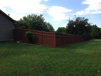 A backyard with a green lawn, a red wooden fence, a house on the left, and trees and shrubs in the background.