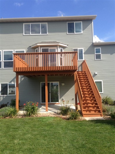 A multi-story house with a red wooden deck and stairs, a green lawn, and a patio area with plants and shrubs.