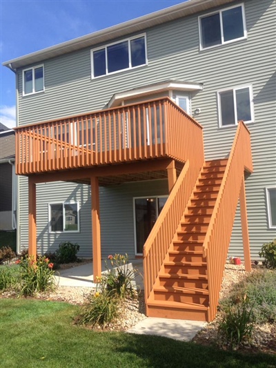 A multi-story house with a red wooden deck and stairs, a well-maintained lawn, and a landscaped area with plants and shrubs beneath the deck.