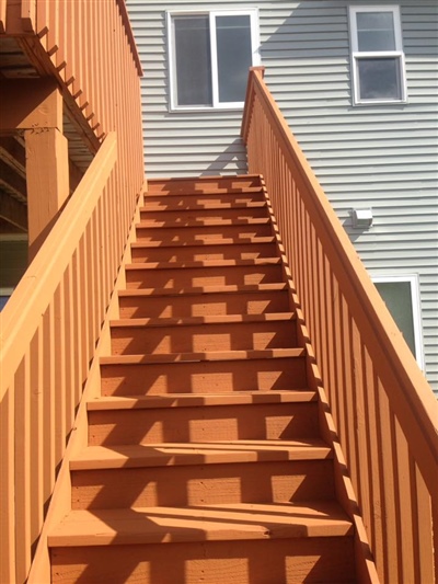 A close-up view of red wooden stairs leading up to a deck on a multi-story house with gray siding.