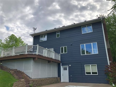 A three-story house with dark blue siding, a large elevated deck, and multiple windows, set against a cloudy sky.