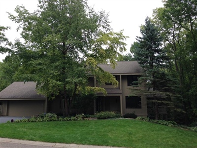 A two-story house with a brown exterior, surrounded by lush green trees and a well-maintained lawn.