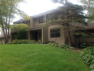 A two-story house with a brown exterior, surrounded by a lush green lawn, trees, and well-maintained shrubs and plants.