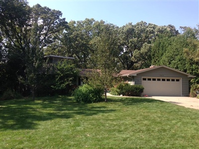 A single-story house with a tan exterior, a two-car garage, a lush green lawn, and surrounded by trees and bushes.