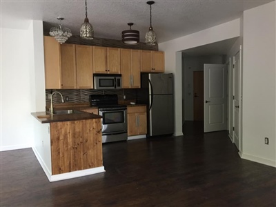 A modern kitchen with wooden cabinets, stainless steel appliances, dark hardwood floors, and pendant lights hanging above the countertop.