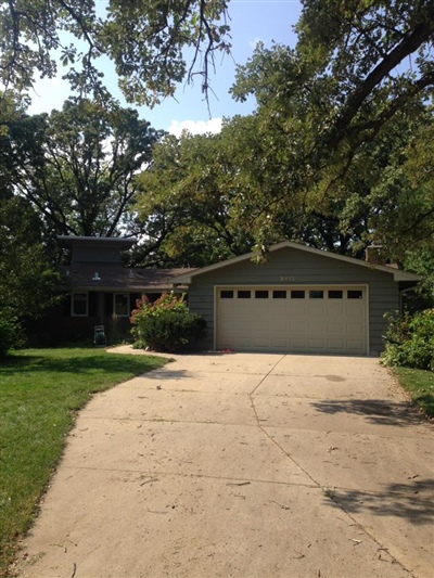 A single-story house with a tan exterior, a two-car garage, a concrete driveway, and surrounded by trees and greenery.