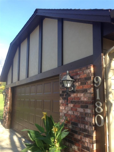 A close-up view of a house with a brown garage door, brick exterior, a wall-mounted light fixture, and the house number "9180" displayed.