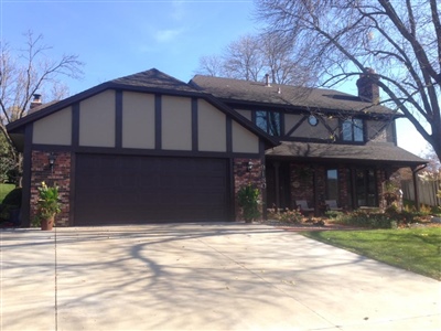 A two-story house with Tudor-style architecture, a brown garage door, brick and stucco exterior, and a large driveway, surrounded by trees.