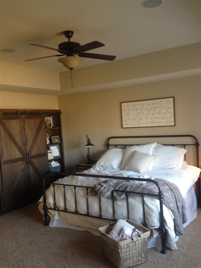 A bedroom with beige walls, a metal bed frame, white bedding, a ceiling fan, a rustic wooden wardrobe, and a wicker basket at the foot of the bed.