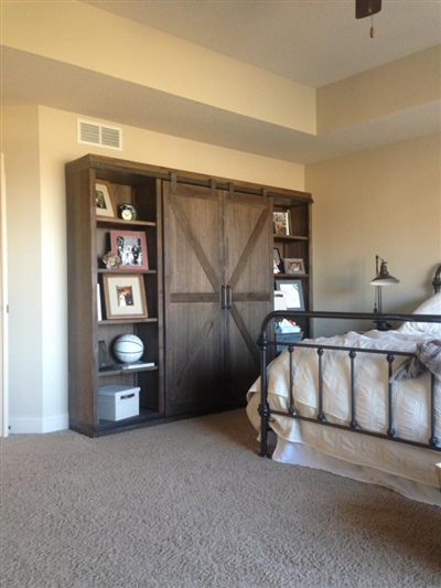 A bedroom with beige walls, a metal bed frame, a rustic wooden wardrobe with sliding barn doors, and carpeted flooring.