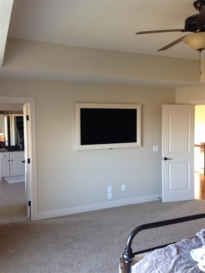 A bedroom with beige walls, a ceiling fan, a wall-mounted TV, an open door leading to a bathroom, and carpeted flooring.