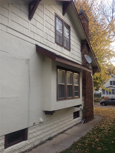 The side view of a house with white exterior walls, brown window frames, a small bay window, and a brick chimney, with fallen leaves on the ground.