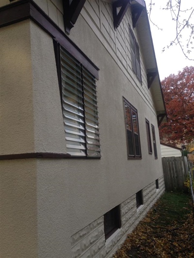 The side view of a house with white exterior walls, brown window frames, and horizontal window blinds, with fallen leaves on the ground and a fence in the background.