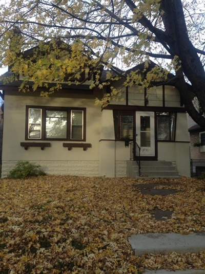 The front view of a house with white exterior walls, brown window frames, a small porch with steps, and a yard covered in fallen leaves, with a large tree in the foreground.