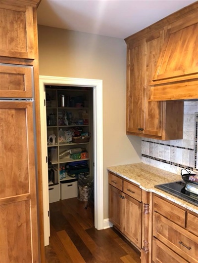 A kitchen with wooden cabinets, granite countertops, and an open pantry door revealing shelves stocked with various items.