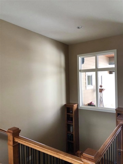 A stairwell with beige walls, wooden handrails, and a window letting in natural light, with a small shelf visible at the bottom of the stairs.