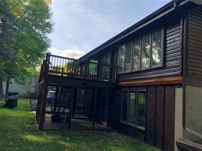 A two-story house with dark wood siding, large windows, and an elevated deck with a staircase, surrounded by green lawn and trees.