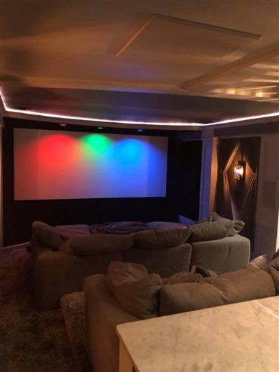 A cozy home theater room with a large screen displaying red, green, and blue lights, surrounded by plush gray sofas and ambient lighting.