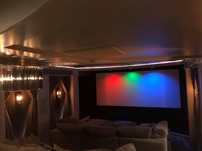 Cozy home theater with plush gray seating, decorative wall sconces, and a large screen displaying red, green, and blue lights.