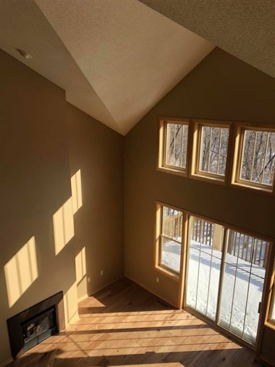 Sunlit, empty room with high vaulted ceilings, large windows, and a sliding glass door leading to a snowy outdoor scene.