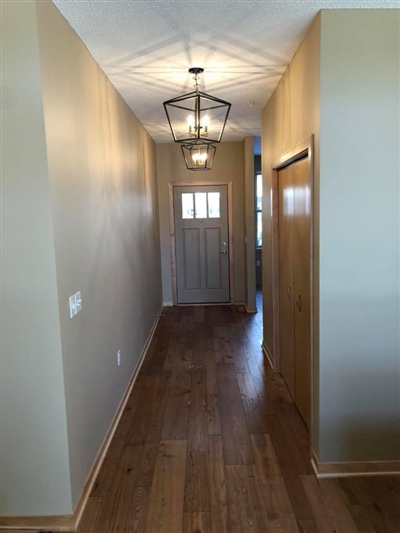 Long hallway with wooden flooring, light beige walls, a front door with a window, and a hanging light fixture.