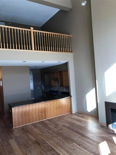 Open living space with wooden flooring, high ceilings, a loft area with a railing, a fireplace, and a kitchen with wooden cabinetry.