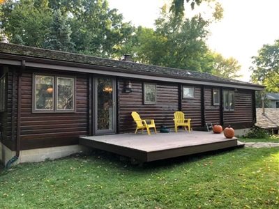 A single-story house with dark wood siding, large windows, and a small deck with two yellow chairs, set in a grassy yard with trees.